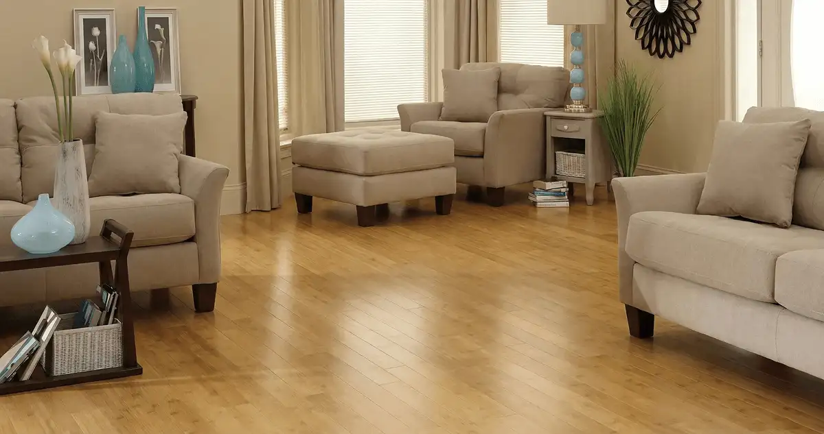 A living room with professionally installed bamboo flooring, beige sofas, a matching ottoman, a side table with a blue lamp, vases, framed pictures, and large windows with blinds and curtains in Worcester, MA