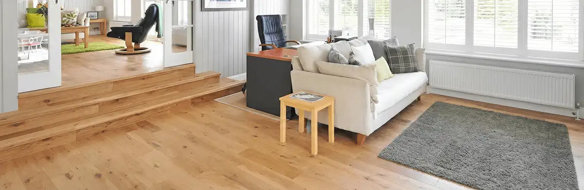 A modern living room with professionally installed laminate flooring, a beige sofa, a small side table, a gray rug, and large windows with white shutters in Worcester, MA